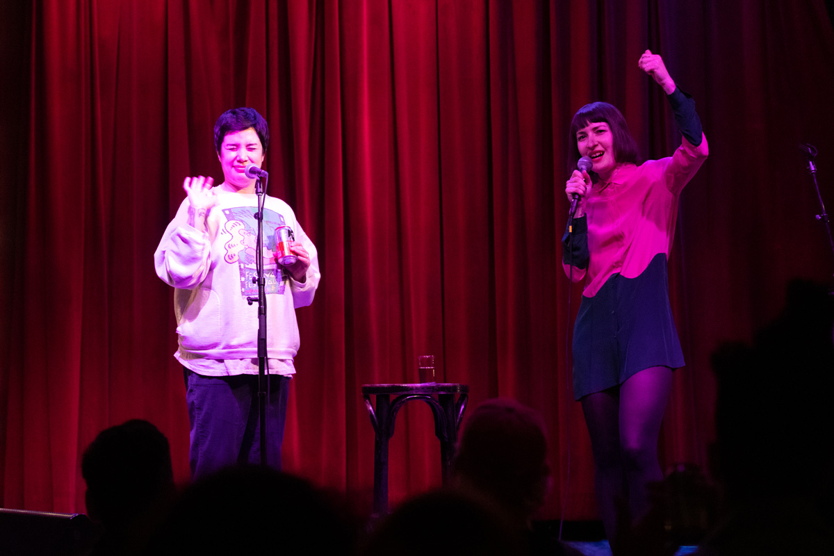 Photo of Clare O'Kane and Emily Panic performing on stage at their monthly show, Windbreaker. Clare is in from of a standing mic and in the middle of telling a joke with her face scrunched up and a can of coke in her hand. Emily is holding a mic and is smiling with one arm raised in a fist. There is a red velvet curtain behind them and a small black stool with a glass of water sitting on it between them. A few of the audience members' heads from the front row are just visible. 