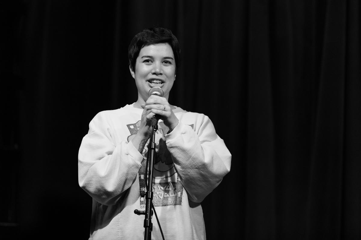 Close up black and white photo of of Clare O'Kane mid-speech with a smile on her face during her and Emily's show Windbreaker. She is holding a mic on a mic stand with both hands in front of her.