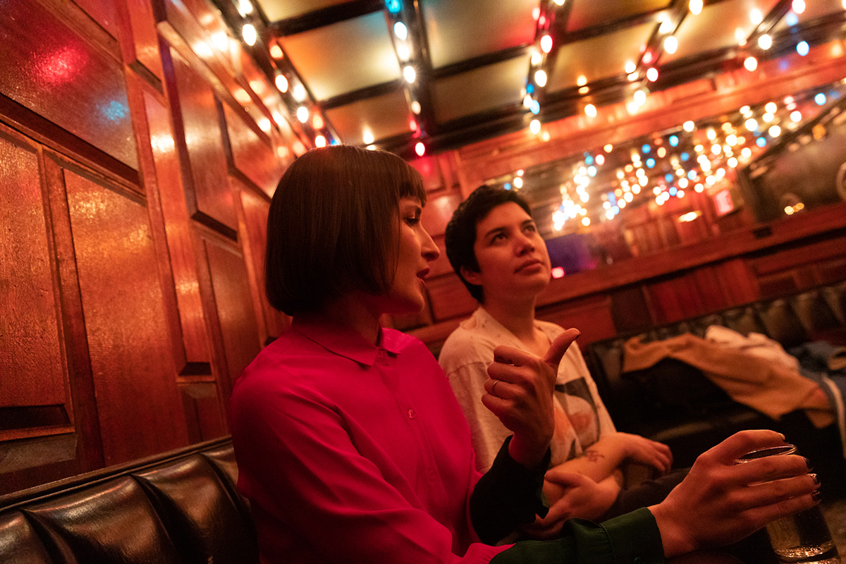 Photo take on Emily Panic and Clare O'Kane sitting in a large booth at a restaurant/bar that is dimly lit except for the strands of string lights running across the long of the ceiling.