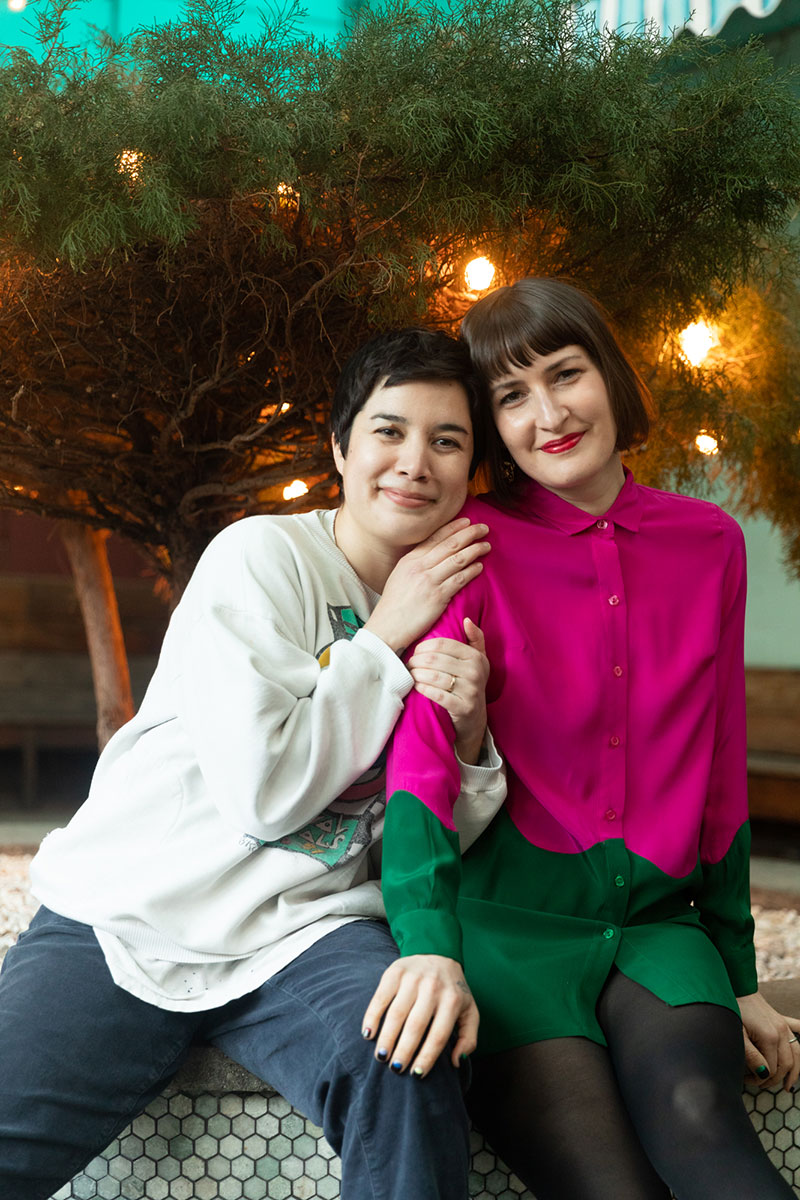 Photo of Clare O'Kane and Emily Panic sitting next to each other on a bench with yellow string lights wrapped around a tree in the background. Clare is hugging Emily's arm and they are both smiling with their heads are tilted together.