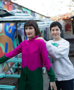 Photo of Emily Panic (left) and Clare O'Kane (right) acting goofy together. Emily has a cropped brown bob with bangs and is wearing a fuchsia pink collared shirt and Clare has a black pixie cut and is wearing a light gray sweatshirt. Emily has her arms spread out in front of her and Clare has her hands on Emily's shoulders.