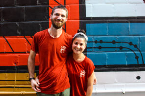 Photo of Maria La Mota and Chason Spencer in their red Chancho King shirts, smiling and posing side-by-side with their arms around each other in front of a red, white and blue art-deco painted brick wall.
