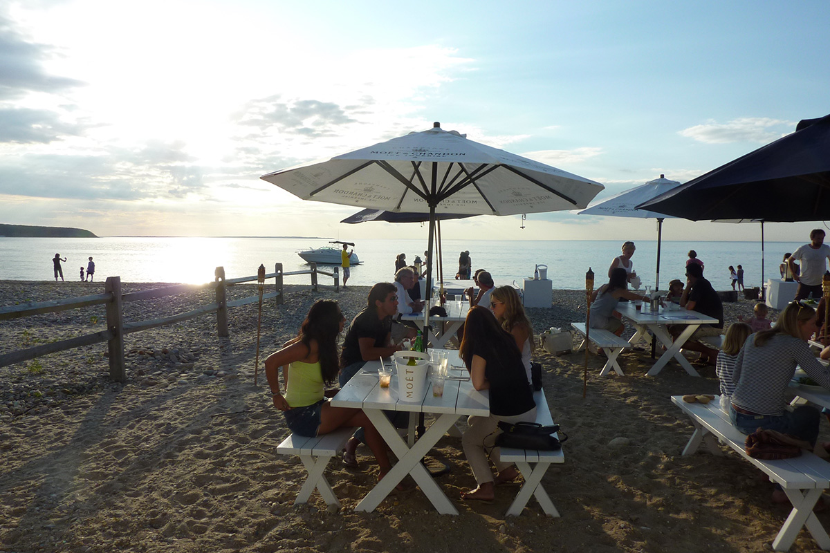 Photo of a group of people eating at the white wooden picnic tables on the sandy shore of Navy Beach. The sun is setting in the background and the water is glass.