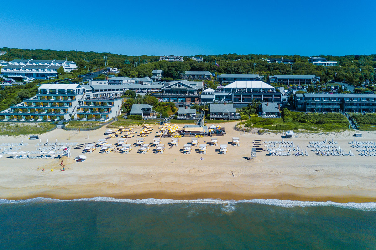 Aerial photo of Gurney's resort in Montauk. The large buildings of the resort have blue accents. Rows of beach chairs and cabanas line the light brown sandy shore in front of the buildings. 