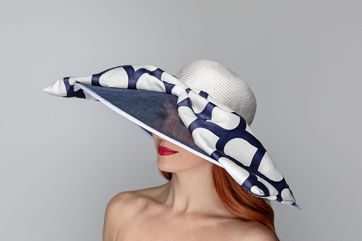 Photo of a red-haired woman wearing a wide-brimmed white and blue Formé Millinery hat. The brim is made of a semi0-transparent navy blue colored material and the base of the hat is white tweed. A large piece of material with a navy blue and white chain-like pattern is pinned in the center around the middle of the hat and look like it is tied in a large knot.