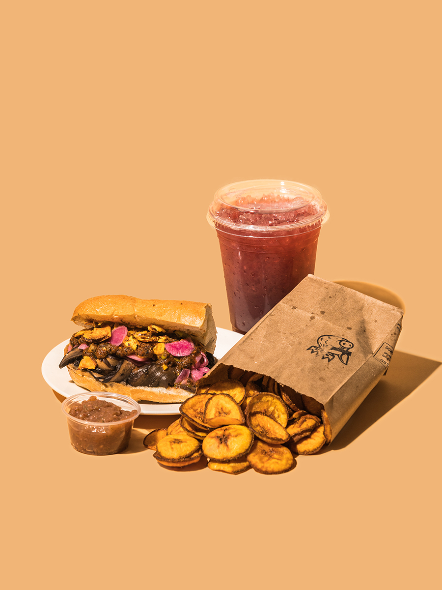 Photo of the original meal served by Chazncho King against a tan background. A half of a sub sandwich with brown meat and sautéed pink and yellow plantain is sitting on a white plate with a small plastic cup of brown sauce in front of it. Next to the sandwich is a paper bag with yellow plantain chips spilling out of it. Behind the paper bag is a plastic cup of light red aqua Fresca.