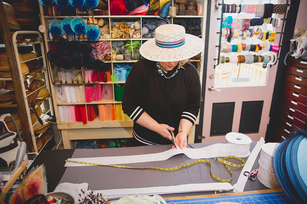 Photo of a Formé Millinery seamstress wearing a white wide-brimmed hat cutting custom sweatbands for the Keeneland Collection. her work shop is stacked from floor to sealing with various colors of ribbon and yarn as well as wooden hat molds.