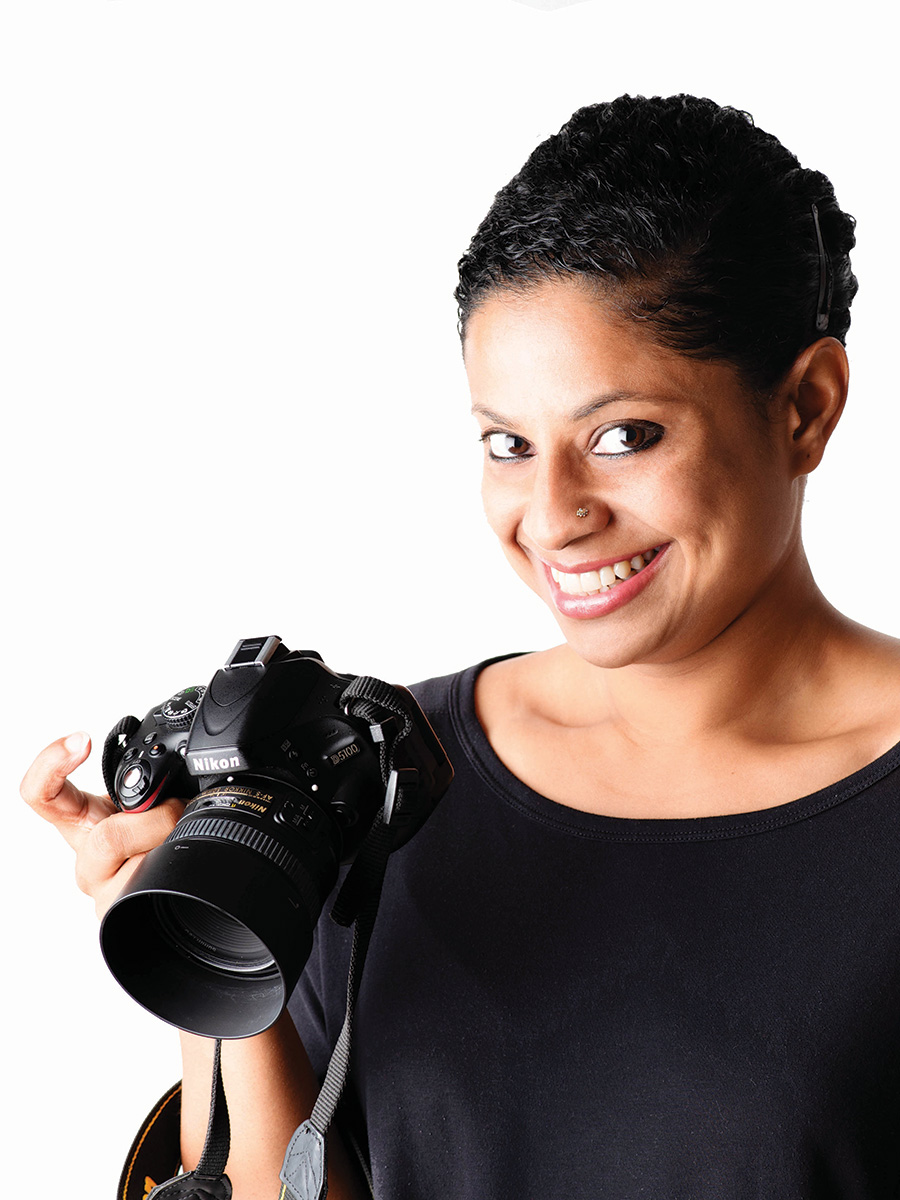 Portrait/head shot of Food Photographer Dyutima Jha taken from a sight upper angle. Dyutima is looking slightly up at the camera and smiling. She is wearing a black shirt and holding a black Nikon camera in her right hand.