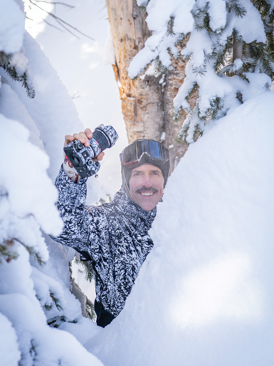 Photo of Winter Photographer Dean Blotto Gray holding his snow-covered camera up from behind some green spruce trees covered in large mounds of snow. Dean is wearing a black ski jacket covered in the white snow and some black ski goggles. He is smiling at the camera. 