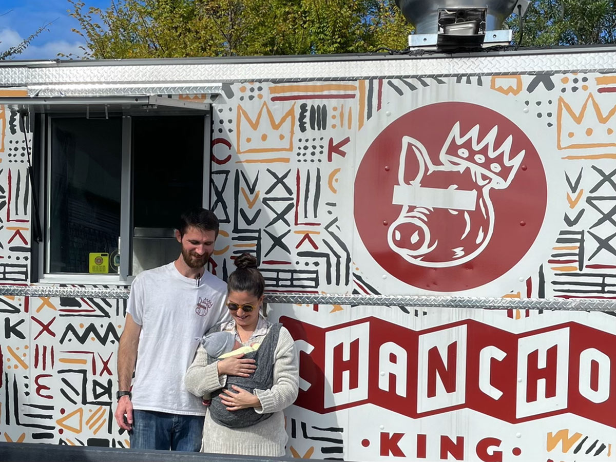 Chason Spencer and Maria La Mona standing in front of their Chancho King food truck. Maria is holding their baby in her arms and both Chason and Maria are looking down and smiling at the baby. The white Chancho King food truck is covered abstractly sketched yellow, black and red shapes and lines. In the center of the truck there is a large red circular logo with the outline of a cartoon pig wearing a crown on one ear. underneath the Chancho King logo are the words "CHANCHO KING".