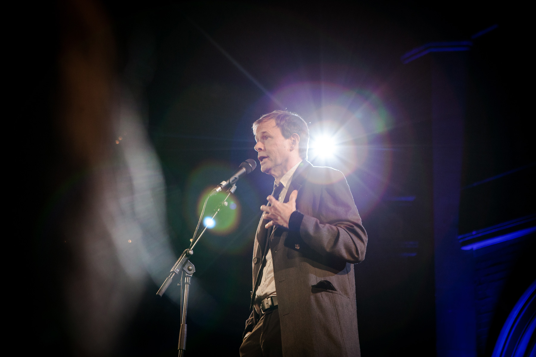 Photo of Author George Dawes Green standing on stage in a brown suit speaking into the microphone as he tells a story to the crowd for The Moth at a live storytelling event. 