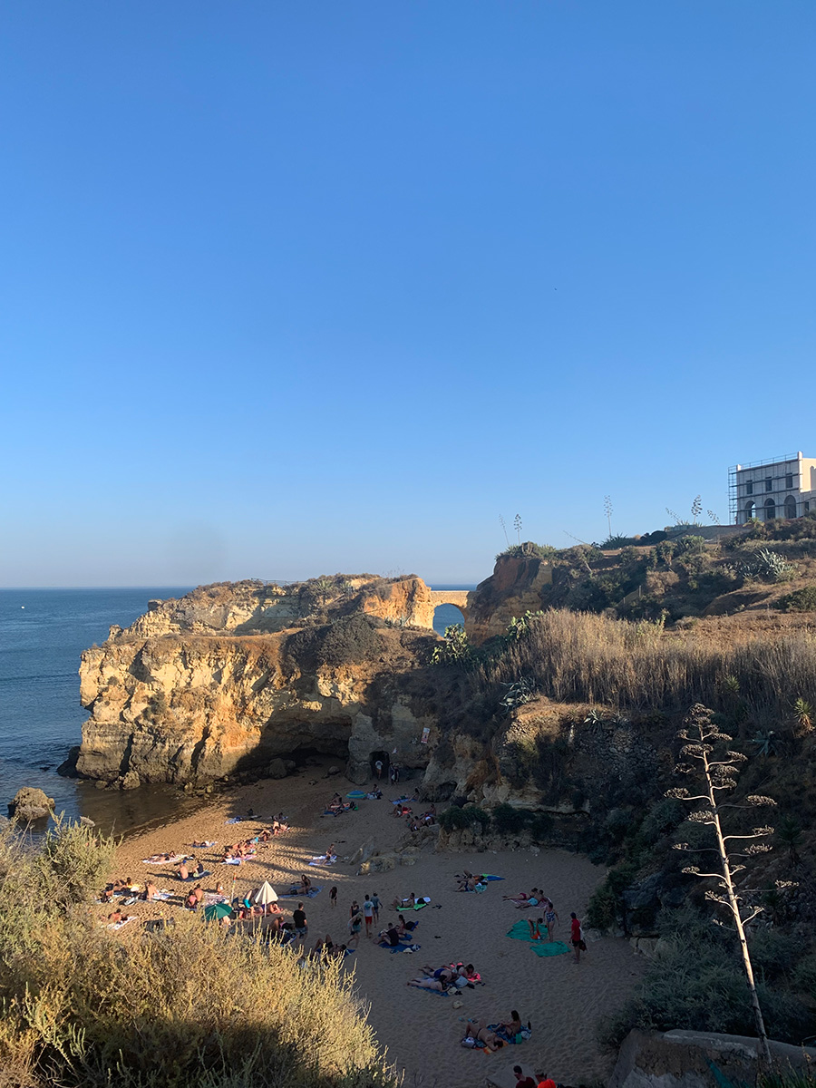 Photo of a rocky, cliff-lined beach cove in Lagos. A group of beach-goers are lounging in the partially shaded sandy are of the cove on towels. 