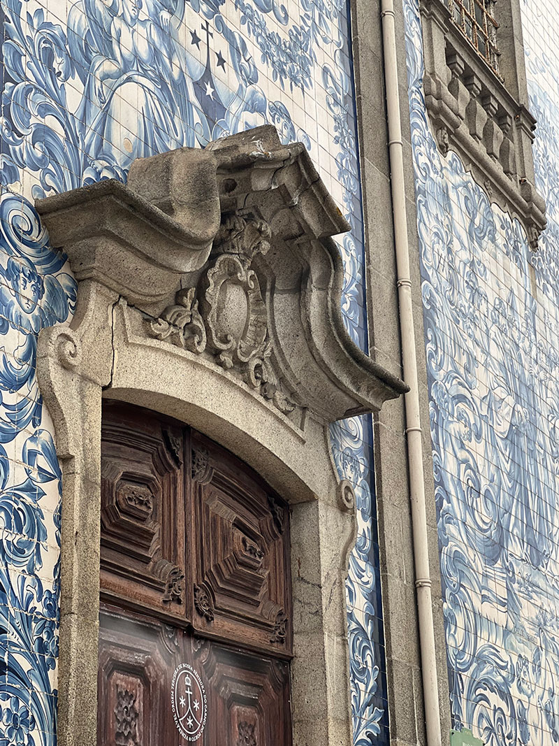 An elaborate stone door frame rests against a wall of blue and white tiles. The door itself is dark wood and carved. 