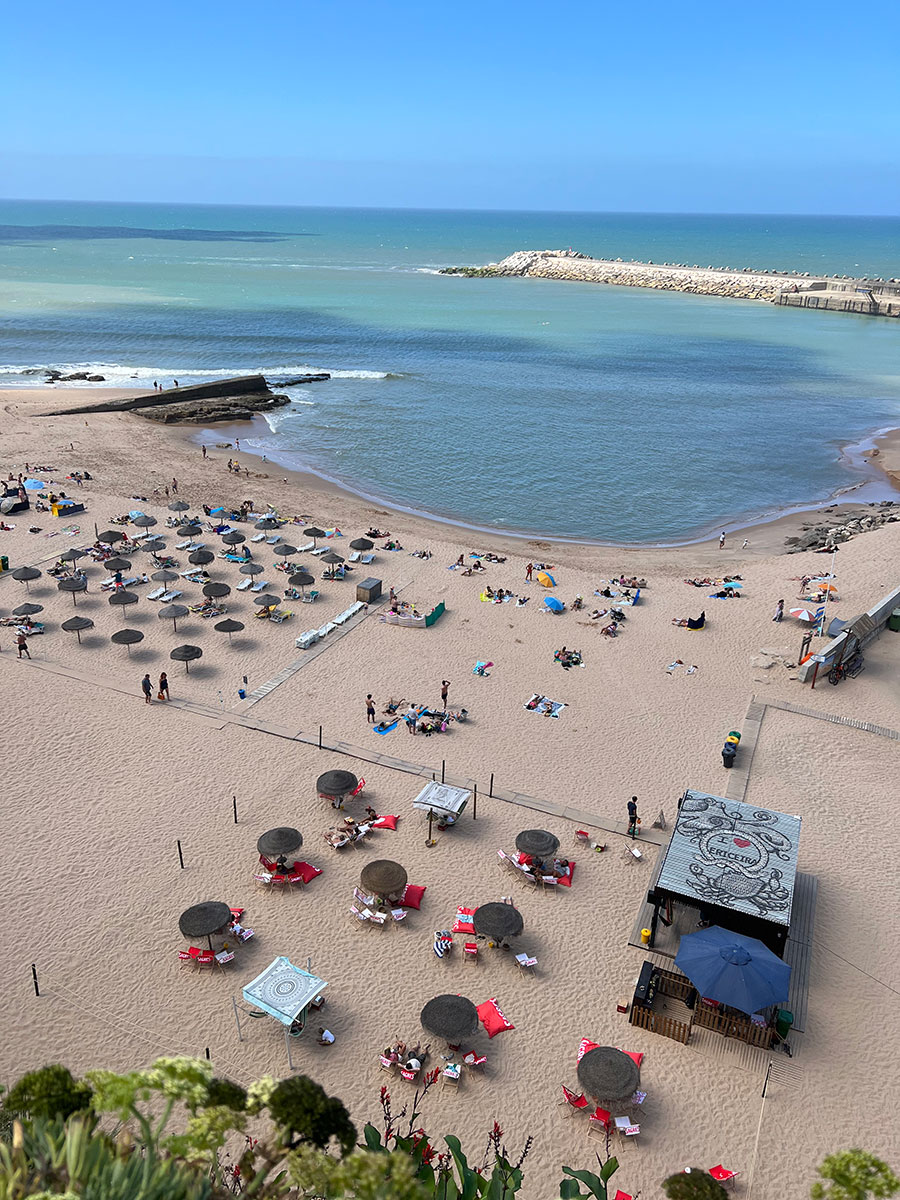 Photo take from above of a beach club in Ericeira. Lines of dark gray umbrella are sectioned off for guests on the sandy beach that butts up to the light blue-green water.