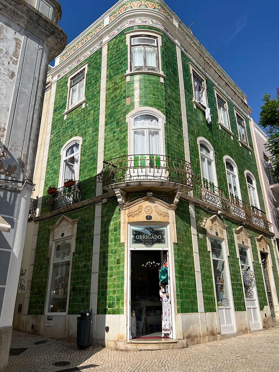 Photo of a tall, emerald green-tiled building on the corner of a tan brick street in Lagos Old Town. Windows with white wooden trim line the outside of the building and are adorned with small cement engravings. The sign on the door reads "Obrigado." 