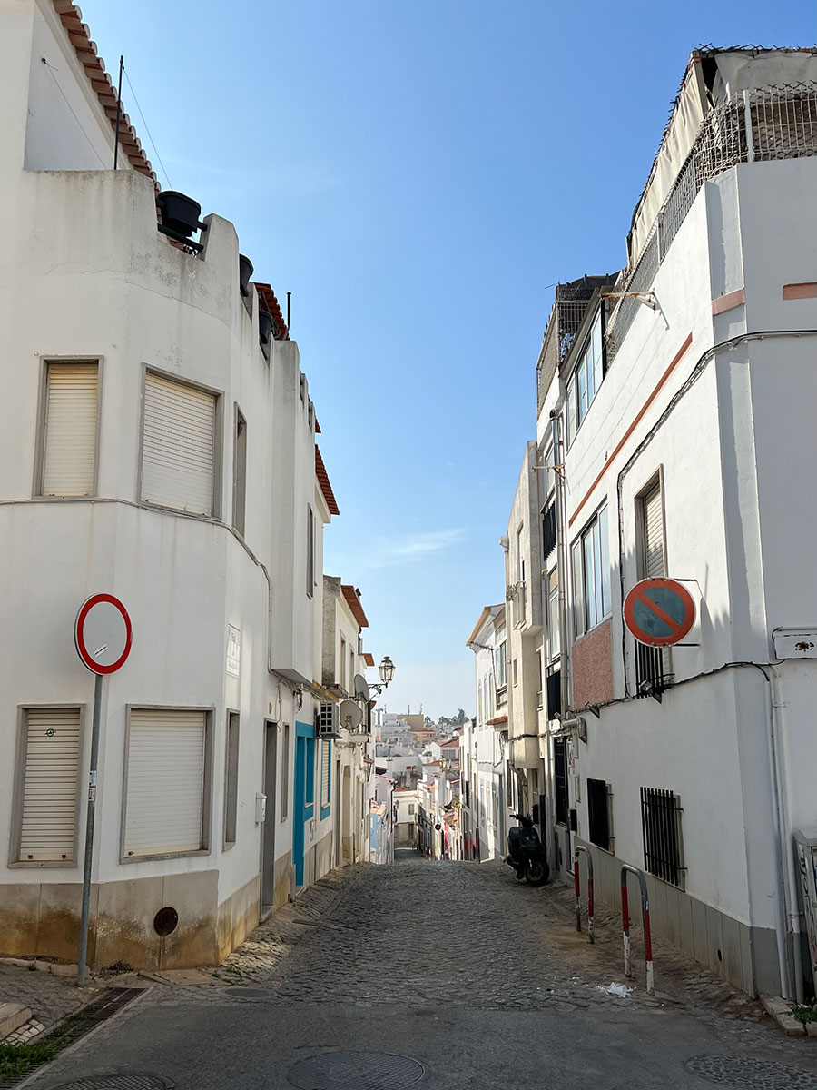 Photo of a narrow, gray cobblestone street in Lisbon, Portugal. The building lining the street are made of white cement.