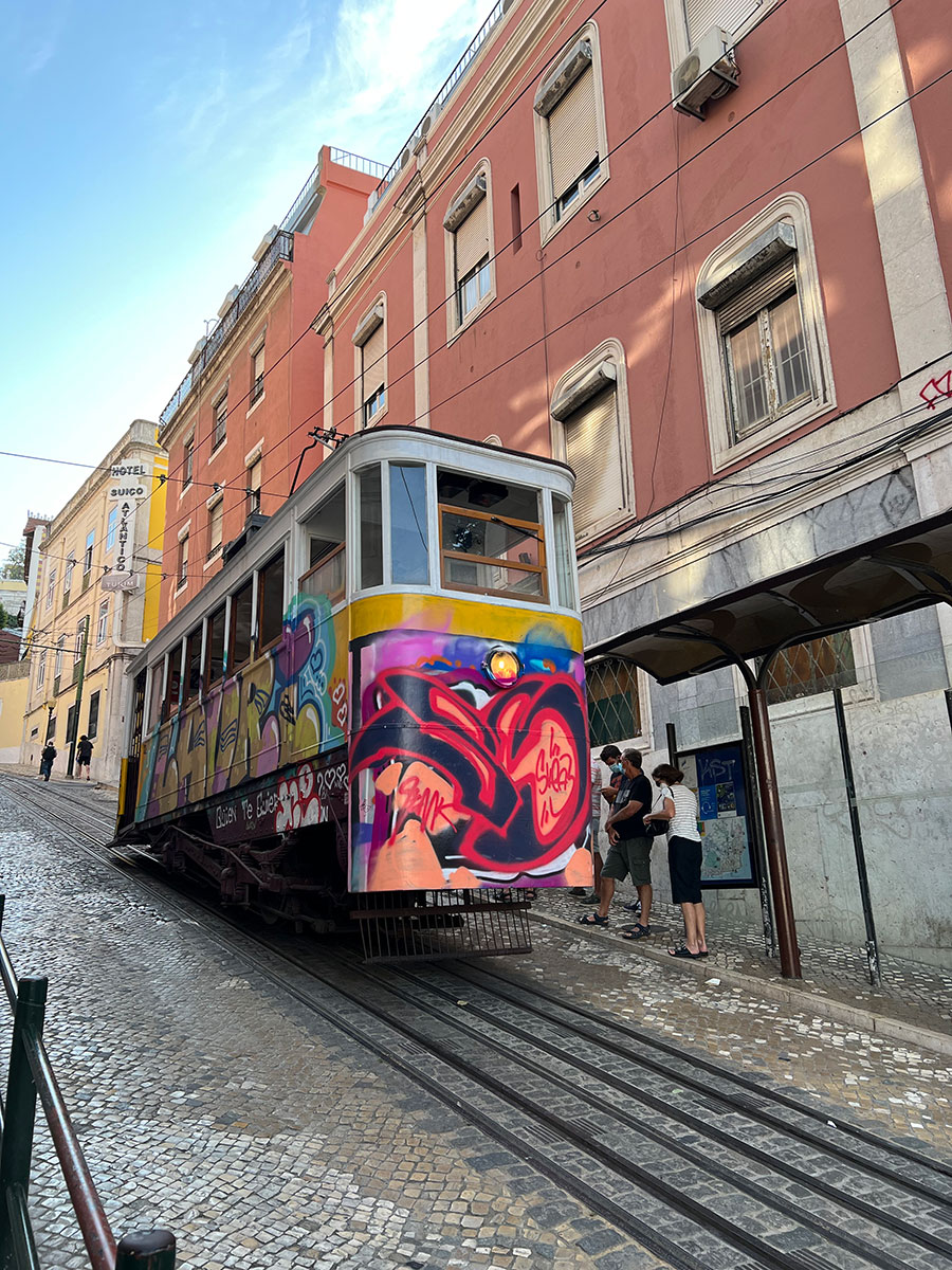 Photo of a yellow tram car in Lisbon covered in decorative bright red and purple graffiti paintings. The tram is going downhill along a narrow cobblestone street lined with orange and yellow buildings.