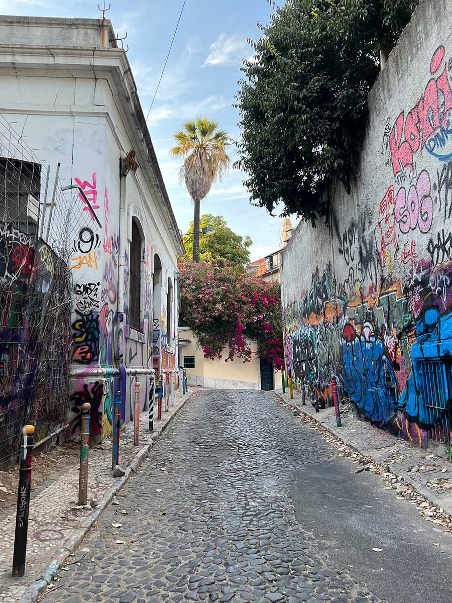 Photo of a small, quaint cobblestone alley in Lisbon lined with small white building covered in decorative, multi-colored graffiti. The building at the end of the alleyway is covered in overgrown lush greenery with bright pink flower blooms