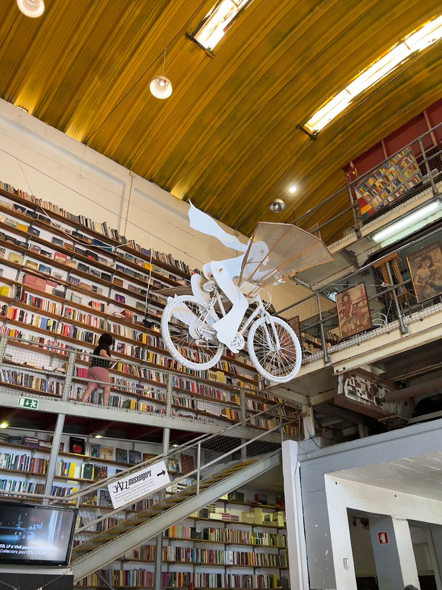 Photo of a bookstore with tall ceilings and multiple floors of walls covered from floor to ceiling in rows of books. A white sculpture go a person in a cape riding a flying bicycle is hanging from the ceiling.