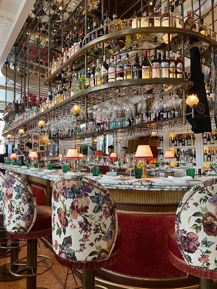 Photo of a highly decorative and fancy restaurant bar. The swivel chairs and the bar itself are covered in white purple and light pink vintage floral upholstery. The bottom go her barm is lined with gold trimming and red velvet panels. Lots of wine glasses hang upside down from the top of the metal grate hanging above the bar and various bottles of alcohol, mixers, wine and more line the shelves of the faded gold grate.