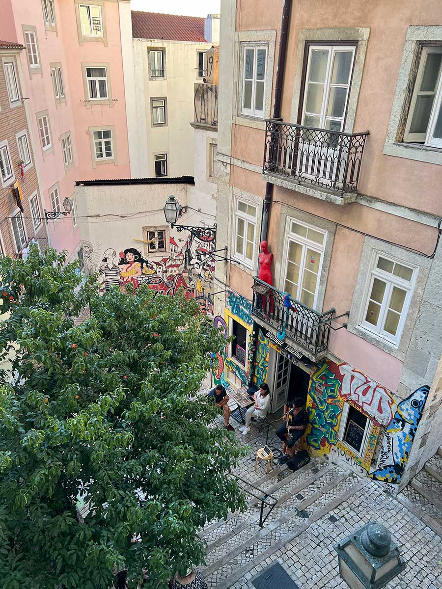 Photo take from a terrace in one of Lisbon's many neighborhoods of the art-covered and graffitied tan cement buildings along a quaint cobblestone alley in Lisbon. A few people are relaxing in chairs and chatting outside the doors of one of the buildings. 