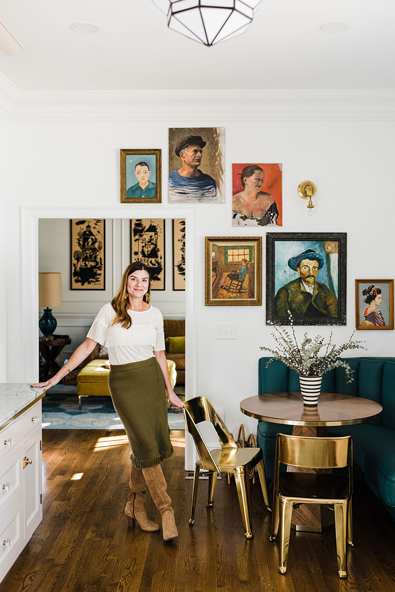 Photo of Natalie Papier standing in the doorway of one of the rooms that she has designed. An array of painted portraits of different people in different styles are hung on the white wall behind her. A dark emerald green booth is in the corner with a golden tabor and chairs in front of it. 