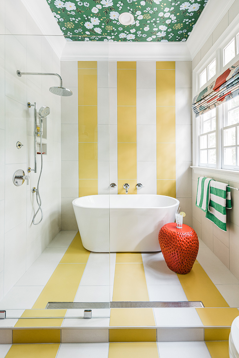 Photo of a bathtub and shower. Alternating yellow and white vertical stripes are running continuously down from the top of the ceiling on the back wall, down the length of the floor and step. The left and right walls are white. There is a window on the right wall with green and white striped towels hanging underneath it and the silver shower head is on the left wall. An oval, white, modern bathtub is pushed up against the back wall. Next to the bathtub there is a bright red strawberry-shaped table with a glass of water on it. The ceiling is crowned and in the center there is a rectangle of green white and yellow floral floral wallpaper. 