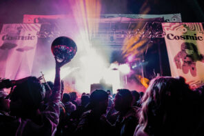 Photo taken from the crowd at a music festival. The large crowd is backlit by the streaming purple, pink and yellow lights illuminating the stage. A person in the crowd is holding up a glimmering disco ball.