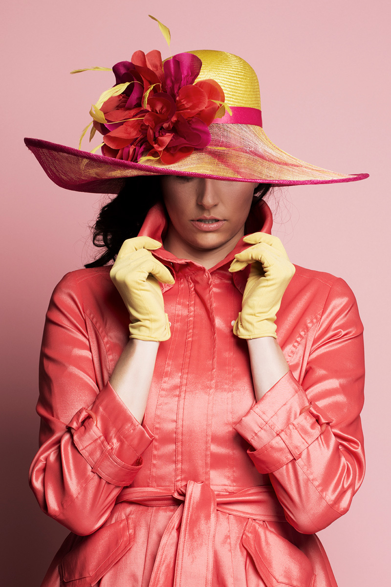 Photo of a woman modeling a Formé Millinery hat against a light pink background. he woman is wearing a shiny pink trench coat and a wide-brimmed yellow hat with hot pink lining and a large pink, orange and yellow flower positioned to the side.