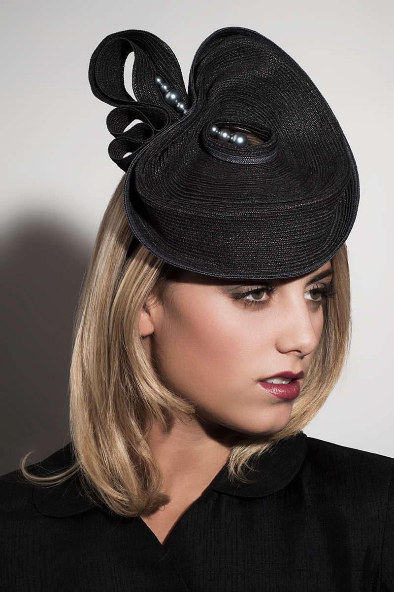 Photo of the profile of a blonde-haired woman modeling a Formé Millinery hat. The hat is black and abstract, almost shaped like a pear. There is a small line of silvery black pearls connecting the large and small pieces of the hat. It is designed to sit just on top of the head and slightly forward.