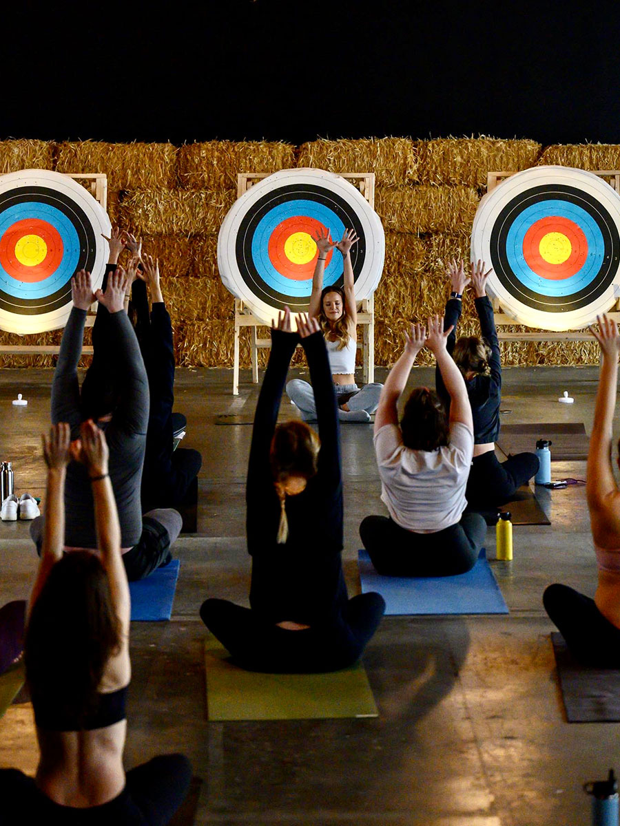 Photo of the back of a group of people sitting cross-legged on yoga mats with their arms reaching up over their heads. There is an instructor sitting at the front of the group performing the same yoga pose while facing the group. Behind the instructor there are 3 large circular targets made of white, black, blue, red and yellow concentric circles standing on wood stands. Behind the targets, there is a wall of stacked golden yellow hay bails.