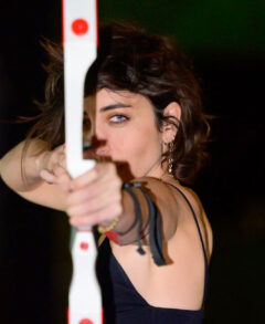 Photo taken close up and straight on in the dark of Kendal Tichner pulling back an arrow against her white and red bow. She is looking directly at the camera down the length of the arrow with a single blue-green eye.
