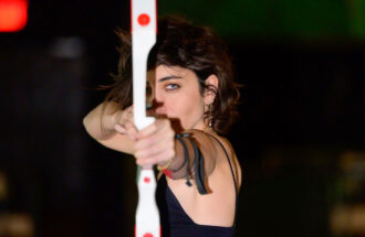 Photo taken close up and straight on in the dark of Kendal Tichner pulling back an arrow against her white and red bow. She is looking directly at the camera down the length of the arrow with a single blue-green eye.
