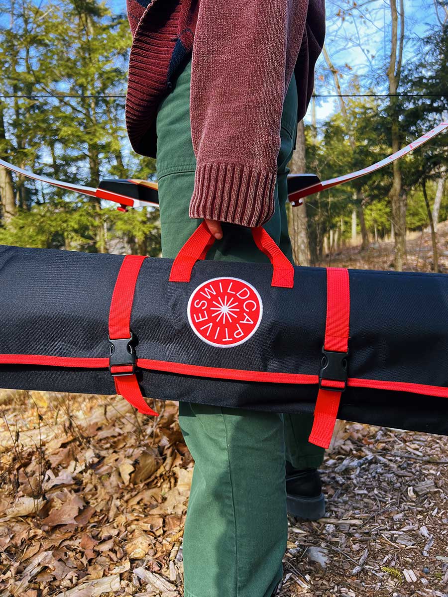 Photo of a person in the woods holding a red and black Wild Captives roll-up case by their side. The case is designed to hold the Wild Captives beginner bow set.