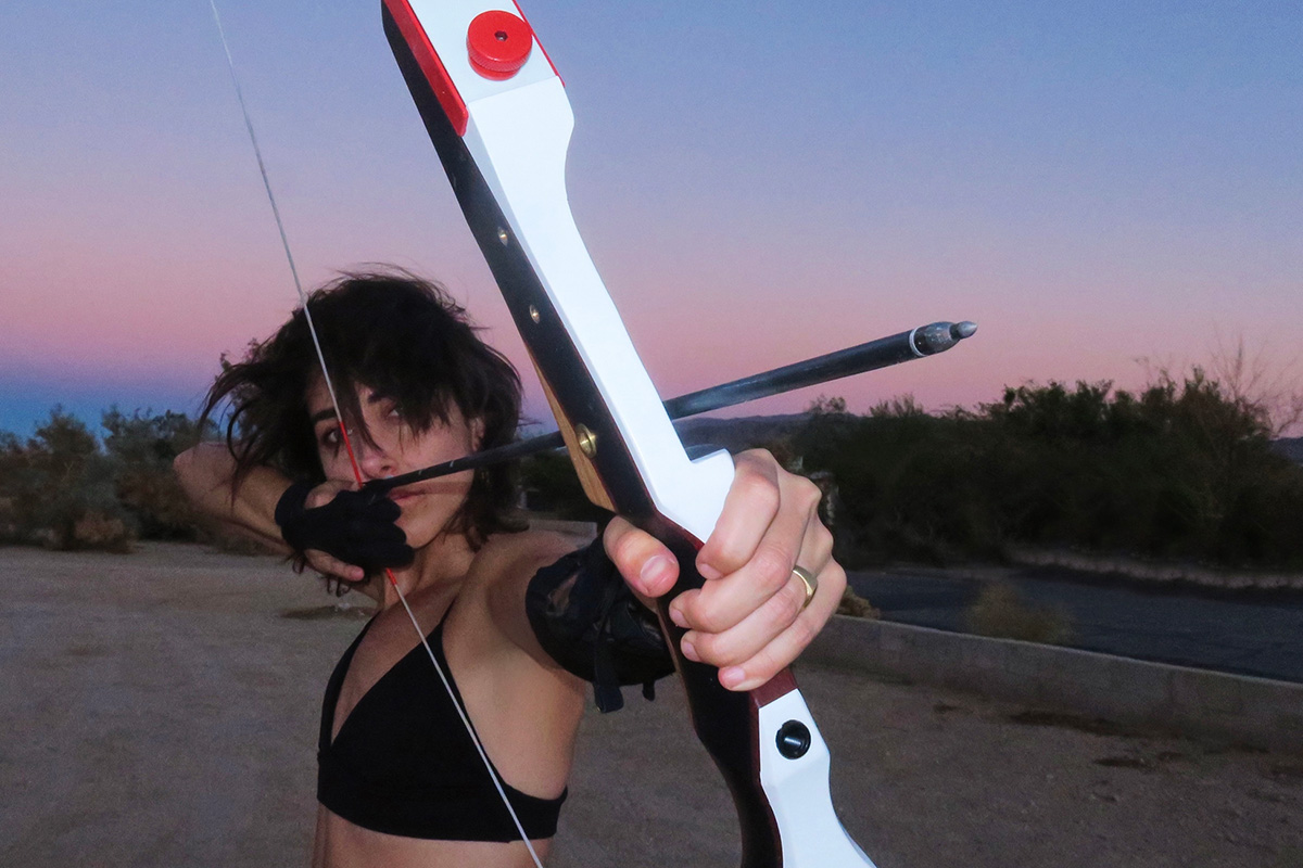 Close up photo taken from the front of Kendall Tichner nocking back a black arrow on her white and red bow while the sun sets behind her, creating a sky full of indigo blue and light pink hues. Kendalls dark hair is windswept and she is wearing a black sports bra as well as black wrist and elbow protectors.
