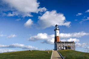 Photo taken from the path leading to The Montauk Lighthouse against a blue sky with a few white clouds.