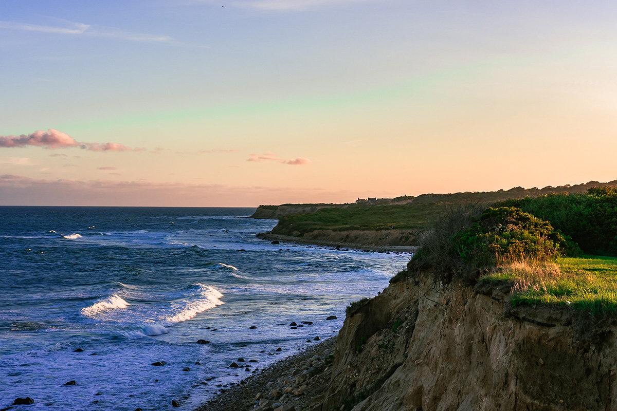 Photo taken from atop a cliff at Ditch Plains Beach. The rocks coast line is topped with patches of green grass and waves from the ocean are lapping up against the cliffs. 