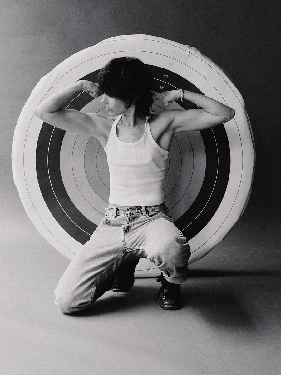 Black and white photo of Kendall Tichner squatting in front of a large target made of concentric circles. Kendall has both of her arms raised and is flexing her biceps while looking to the side.