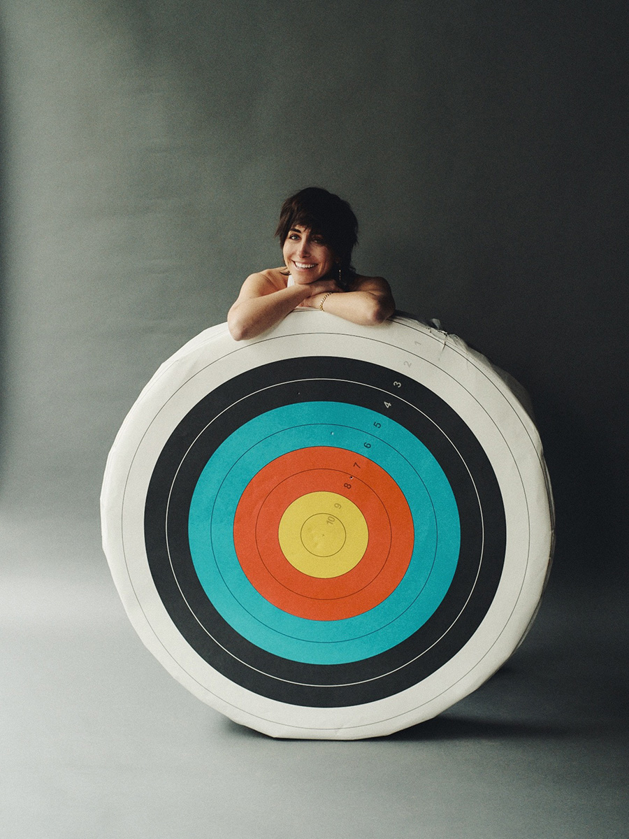 Photo of Kendall Tichner standing behind a large target with white, black, blue, red and yellow concentric circles against a gray background and floor. Her body is completely covered by the target except for her arms which are crossed and resting on the top of her target, and her head which is resting on her folded hands while she smiles at the camera.