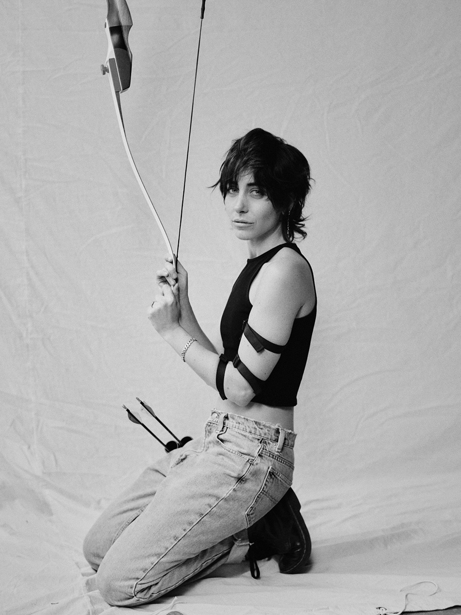 Black and white photo of Kendall Tichner kneeling down while holding her bow up in the air. A quiver of arrows is leaning against her legs. She is looking at the camera.