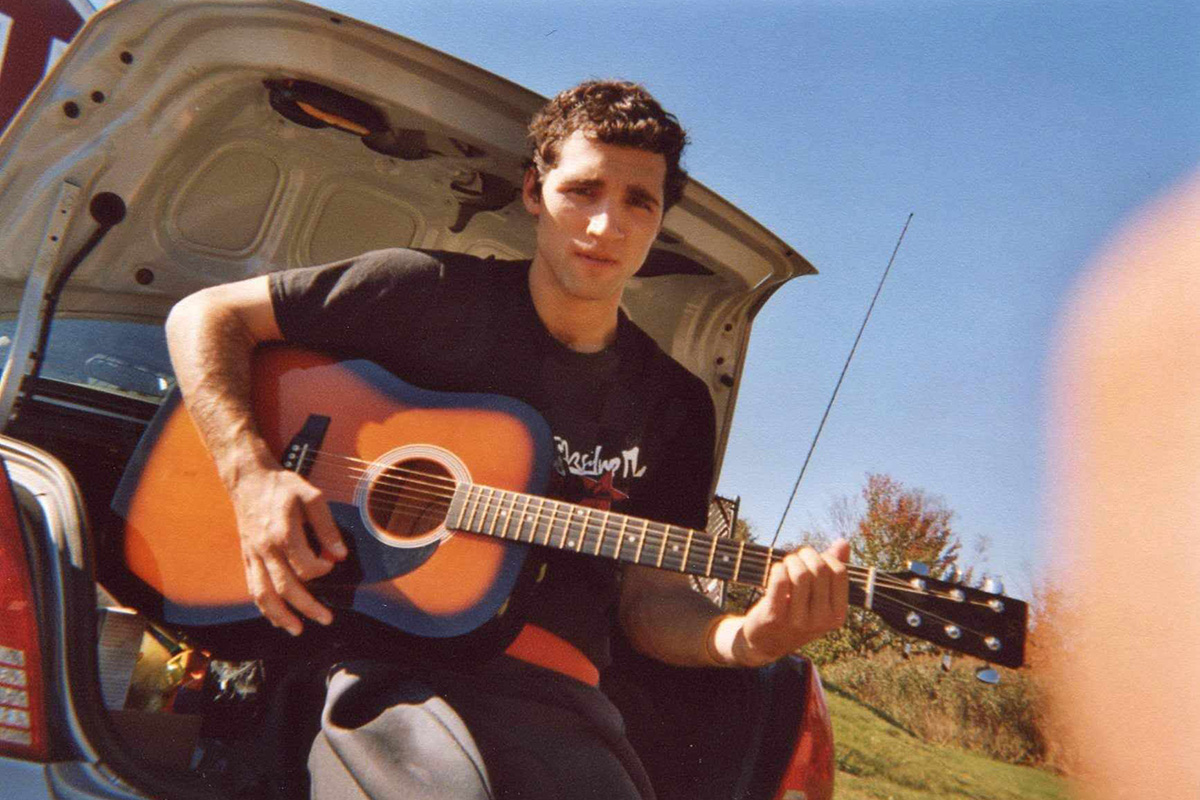 Photo of Hassan Laramée before his accident sitting in the open trunk of a car playing a brown and black guitar.