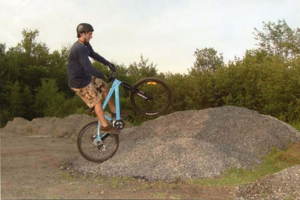Photo of Hassan Laramée doing a wheelie on a light blue bike off of a mound of gravel.