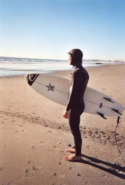 Photo of Hassan Laramée before his mountain biking accident standing on a beach in a black winter wetsuit with a surfboard under his arm looking out at the ocean.