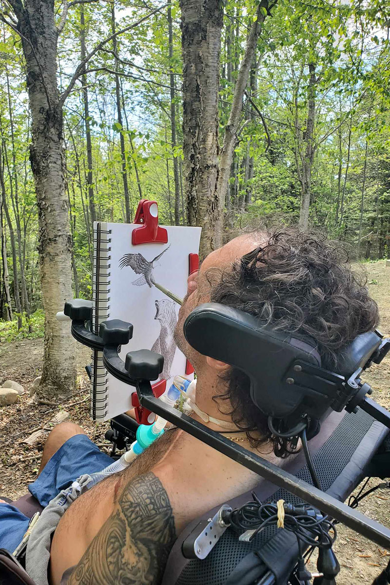Photo of Hassan Laramée drawing a photo. Hassan is sitting in a wheelchair in a green wooded forest drawing in a sketchbook that is propped up with clips and a metal arm that extends from his wheelchair. He is holding a pen in his mouth and using it to draw.