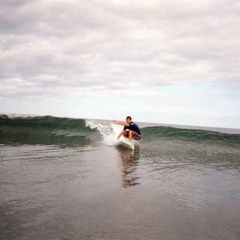 Photo of Hassan Laramée before his accident surfing a small wave in the ocean. Hassan is crouched down on his surfboard and gripping its edge as he surfs toward the camera.