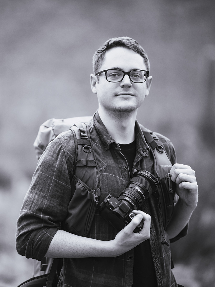 Black and white portrait of Landscape/Astrophotographer Sean Parker standing outside. Sean is wearing a hiking backpack with a large black camera strapped onto the front across his chest. He is wearing glasses and a flannel.