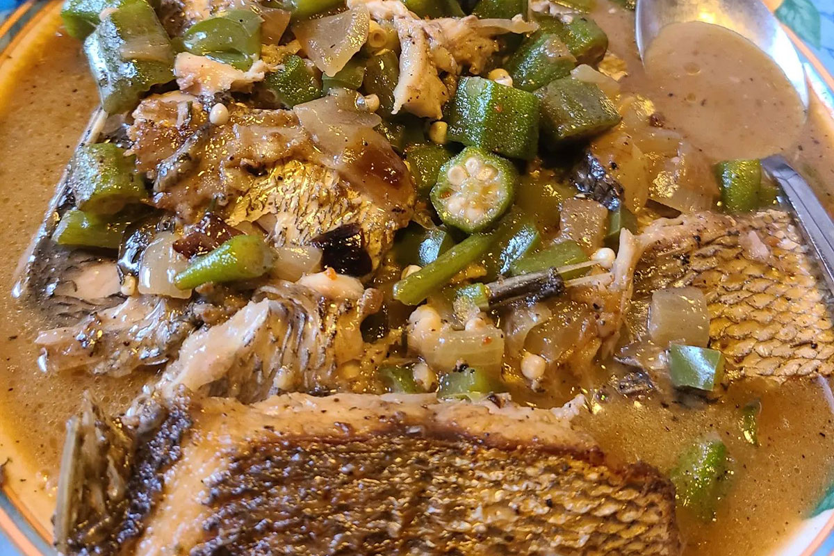 Photo of a bowl of fish stew. The broth is a light yellow-brown color. The dish consists mainly of sliced up bits of green okra and translucent yellow onions spooned over top of large chunks of grilled fish with the skin still on it.