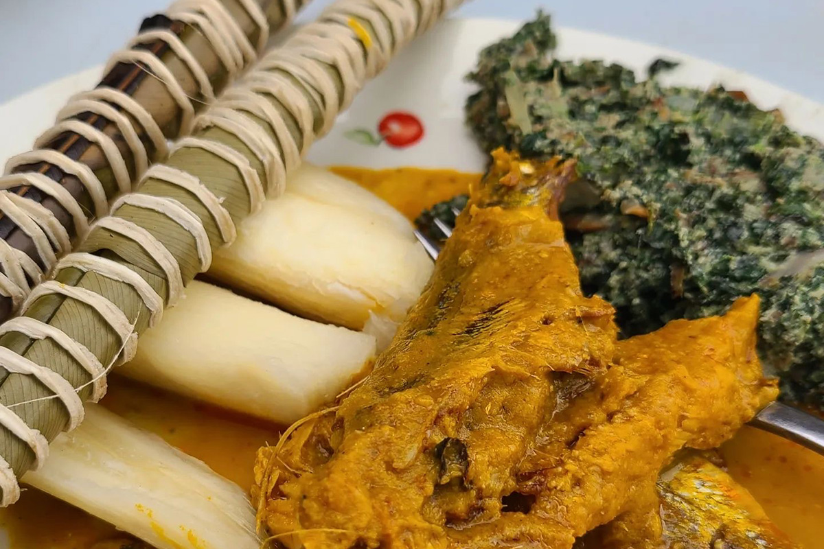 Photo of a soup native to Cameroon. The two light green sticks sitting on top of a light yellow vegetable are pieces of cassava rooted steamed and wrapped in banana leaves. Next to them there is a piece of orange-yellow smoked fish. Behind the fish is a pile of steamed dark green vegetables. The soup broth is a light orange.