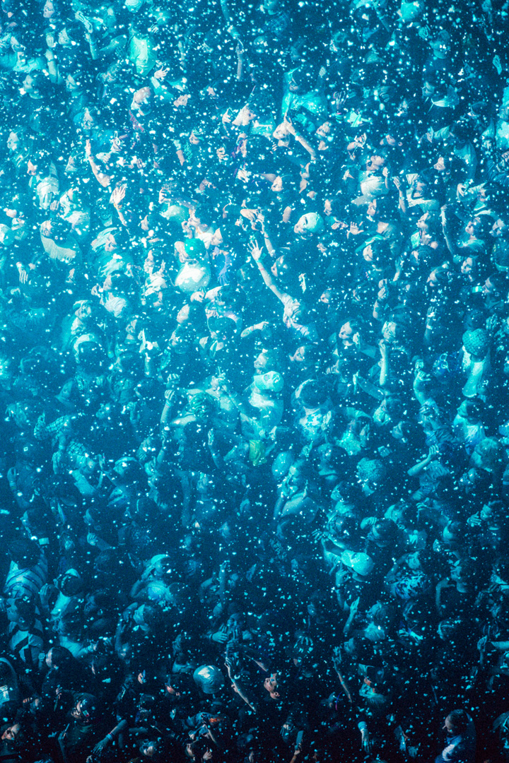 Arial view of a crowd standing around close to another. Some of them have their arms raised in the air, others are clapping, and some are holding up cameras. There is confetti falling on the crowd and there are blue lights shining on everyone giving the image a blue tint.