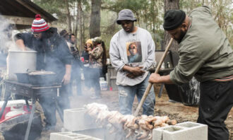 Photo of BJ Dennis helping roast a row of whole chickens skewered on a large spit at the Gulla Geechee community outdoor barbecue.